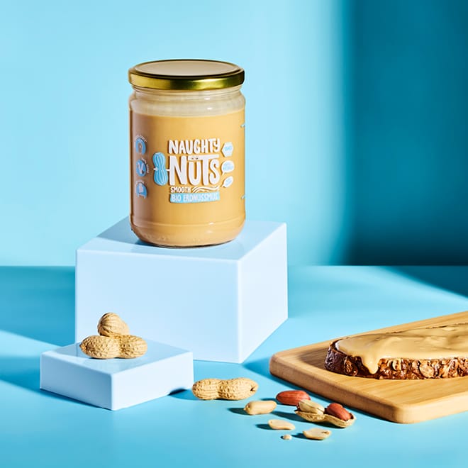 Bio Erdnussmus Smooth (500g Glas) auf einem hellblauen Würfel, Erdnüsse und Brot mit Erdnussmus