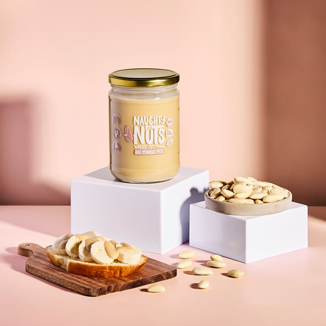 Bio Weißes Mandelmus (500g Glas) auf weißem Podest, daneben Mandeln und Bananenbrot